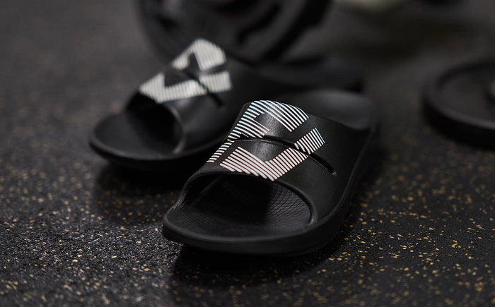 FJ X OOFOS Men's OOahh Sport Slide Sandal - Black close up of shoes on gym floor | lifestyle