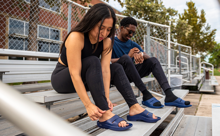 Men's OOahh Slide Sandal - Navy male and female model wearing shoes sitting in bleachers | lifestyle