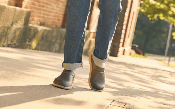 Person wearing gray shoes and blue jeans walking on a sidewalk.