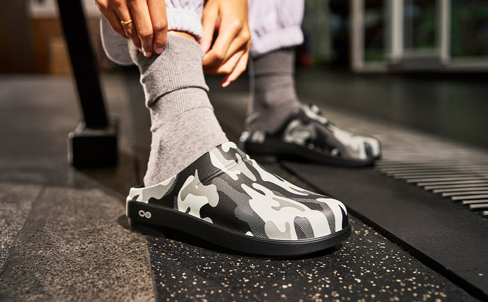 A person wearing gray socks and black-and-white camouflage slip-on clogs in a gym setting, with one shoe being adjusted and the other visible in the background | lifestyle